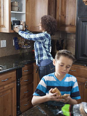 Woman putting pill bottle in locking box in kitchen cabinet while boy makes sandwich.